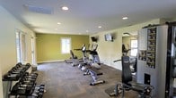 This single-floor fitness center offers a practical and well-organized space designed to support a range of workout routines. The room features neutral walls accented by olive-green paint, creating an energizing atmosphere suited for exercise. Recessed ceiling lights provide consistent, even illumination throughout the space, complemented by natural light filtering in through windows on multiple walls.

The fitness area is equipped with a selection of cardio machines, including ellipticals, a stationary bike, and a rowing machine, arranged to allow comfortable movement between equipment. A wall-mounted strength training station is positioned along one side of the room, offering additional exercise options. Two wall-mounted flat-screen televisions are installed for entertainment during workouts.

A large floor-to-ceiling mirror is affixed to one wall, enhancing the sense of space while allowing users to monitor their form. The flooring consists of durable, patterned carpet tiles, providing cushioning and traction underfoot. Free weights are also visible near the floor, rounding out the available equipment. The overall layout is clean and functional, maximizing usability within the available footprint.