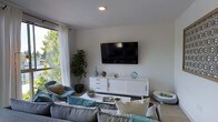 This single-floor space features a thoughtfully designed living area that balances modern aesthetics with comfortable functionality. The living room is anchored by a wall-mounted flat-screen television positioned above a sleek white media console, which provides ample storage through a combination of drawers and sliding cabinet doors. The console is styled with decorative accents, including blue ceramic vases and sculptural objects that complement the room's cool-toned palette.

Seating is generously arranged, with a large, plush sectional sofa in neutral gray paired with teal and cream throw pillows, creating a cozy and inviting atmosphere. A rounded blue accent chair adds a subtle design contrast in the corner near the window. A glass-topped coffee table sits at the center, dressed with a terrarium and reading materials.

Natural light fills the room through a large window dressed with sheer white curtains, offering views of lush greenery outside. Recessed ceiling lighting supplements the natural light, ensuring the space remains well-lit. A tall indoor plant near the window adds a natural, organic element to the otherwise clean and contemporary interior.