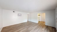 This generously sized 2-bedroom, 1-bathroom single-story residence offers a clean, functional layout designed to maximize livable space. The open living area is a standout feature, showcasing warm wood-look vinyl flooring, crisp white walls, and a textured ceiling that adds subtle visual interest. A large window at the front of the living space floods the room with natural light, creating a bright and welcoming atmosphere.

The galley-style kitchen runs along a single corridor and is efficiently outfitted with white cabinetry, white countertops, and white appliances including an electric range and full-size refrigerator. The wood-look flooring continues here, maintaining a cohesive aesthetic throughout the common areas.

Both bedrooms offer comfortable proportions, each featuring plush neutral carpeting and ceiling fans with integrated light fixtures for added comfort and functionality. The rooms are finished with white walls and clean trim, providing a versatile backdrop for various furnishing styles.

The bathroom is finished in an all-white palette, featuring a vanity with a large mirror, a well-lit fixture above, and ample counter space. The overall design of this unit emphasizes simplicity, cleanliness, and practicality throughout.