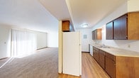 This single-floor space features an open-concept design that seamlessly connects the kitchen and living area. The kitchen is equipped with dark brown cabinetry providing ample storage, a double-basin sink, a full-size refrigerator, and a dishwasher. The countertops offer functional workspace, while the hardwood-style flooring adds warmth and durability to the kitchen zone. A partial wall with upper cabinetry serves as a natural divider between the kitchen and the adjacent living area, creating defined zones while maintaining an open feel. The living area transitions to soft carpeting, offering a comfortable and inviting atmosphere. Vertical blinds on the windows allow for adjustable natural light throughout the space. Textured ceilings and flush-mount lighting fixtures are present throughout, providing consistent illumination. The neutral color palette of whites and beiges creates a clean, versatile backdrop suitable for a variety of interior styles. Overall, the layout is practical and efficient, making good use of the available space across the single floor.
