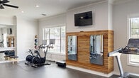 Located in Fort Worth, TX, this fitness center offers a thoughtfully designed workout space that balances functionality with aesthetic appeal. The single-floor layout provides generous open floor space, accommodating a comprehensive range of exercise equipment without feeling cramped.

The interior features crisp white walls accented by crown molding and a striking natural wood slat wall, which serves as a focal point and adds warmth to the space. A decorative textured accent wall with an equestrian mural motif provides an additional design element, framed with gold trim detailing. Multiple wall-mounted flat-screen TVs are positioned throughout for viewing during workouts.

Cardio equipment includes several treadmills and stationary bikes, while the strength training area is outfitted with cable machines, a multi-functional weight rack, a squat rack, and an elliptical. A medicine ball rack and foam rollers are also available for functional training and recovery. Large windows along the perimeter flood the space with natural light and offer views of the outdoor surroundings, including a pool area. Dual ceiling fans ensure consistent airflow throughout. Recessed lighting complements the natural light, and durable dark rubber flooring covers the entire workout area, providing both safety and easy maintenance.