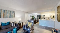 This single-floor residence offers a thoughtfully designed open-concept layout that seamlessly connects the living, dining, and kitchen areas. The living room features light wood-look flooring, a sliding glass door that opens to a private balcony with outdoor views, and generous natural light. A patterned accent chair and contemporary furnishings contribute to the space's modern, stylish ambiance.

The kitchen is equipped with stainless steel appliances including a range, microwave, and refrigerator, complemented by white cabinetry and a light-toned quartz countertop with an undermount sink. An in-unit washer and dryer are conveniently located nearby. The adjacent dining area features a glass-top table beneath a multi-pendant chandelier, with a decorative screen partition adding visual interest.

The primary bedroom includes carpet flooring, a ceiling fan with light fixture, and a well-sized window for natural light. The bathroom offers a quartz vanity countertop, a large mirror, and a tub-shower combination. A flexible secondary room with carpet flooring functions effectively as a home office or additional sleeping space. Throughout the residence, textured ceilings, neutral gray walls, and consistent design choices create a cohesive and comfortable living environment.