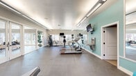 This fitness center in Vacaville, CA offers a thoughtfully designed single-level workout space with a generous open floor plan. The interior features durable rubber and concrete-style flooring throughout, smooth white ceilings with linear fluorescent lighting, and a neutral gray and mint-green color scheme that creates a clean, energizing atmosphere.

The cardio zone is anchored by multiple Star Trac treadmills and upright cycling machines, each equipped with individual mounted monitors for entertainment during workouts. A multi-station cable machine and additional resistance equipment are positioned along one side, providing a comprehensive strength-training option. Wall-mounted flat-screen televisions are installed at multiple viewing angles throughout the space.

A dedicated free weights area features a tiered dumbbell rack, an adjustable weight bench, TRX suspension straps mounted overhead, and a medicine ball collection in assorted weights. Floor mats and stability balls support stretching and functional training routines. Full-length mirrors line key walls, enhancing the sense of spaciousness and allowing users to monitor their form.

Large sliding glass doors and multiple windows along the perimeter allow abundant natural light to fill the space and provide views of surrounding outdoor greenery. A wall-mounted water fountain is conveniently accessible near the main exercise area.