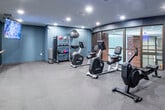 Located at 401 Williams Ave SW in Huntsville, Alabama, this fitness center offers a well-appointed, single-floor workout facility designed to support a variety of exercise routines. The space features a modern aesthetic with dark gray walls, recessed lighting, and durable carpet flooring throughout, creating a focused and energizing environment.

The main exercise area is equipped with Life Fitness cardio machines, including upright and recumbent cycles and a rowing machine, providing a comprehensive range of low-impact cardio options. A dedicated free weights and accessories station holds kettlebells, medicine balls, and stability balls on a multi-tier rack, accommodating strength and functional training needs.

A full-length mirrored wall runs along one side of the room and is complemented by a mounted ballet barre, making the space suitable for stretching, yoga, or barre-style workouts. Large flat-screen televisions are mounted throughout the room, allowing users to stay entertained or follow guided workouts during their sessions.

Floor-to-ceiling glass panels along one wall offer views into an adjacent interior corridor and exposed brick architectural elements, adding a sense of openness to the space. Safety equipment, including an AED and emergency pull station, is clearly mounted and accessible. The fitness center is well-lit with recessed ceiling fixtures and benefits from a clean, organized layout that maximizes usability.