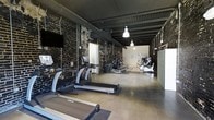 This single-floor fitness center combines a rugged industrial aesthetic with a comprehensive selection of workout equipment, creating a functional and visually striking environment for exercise. The space is defined by its dramatic dark-painted exposed brick walls, which lend a raw, urban character throughout. The ceiling features exposed structural elements finished in dark tones, with industrial-style pendant lighting that illuminates the workout floor effectively.

The main floor is a long, open layout that accommodates a generous lineup of cardio machines, including treadmills and ellipticals from reputable brands such as True Fitness, arranged in a row along one side of the space. Rubber flooring in the free weights area provides safety and durability, supporting a full rack of dumbbells spanning a wide range of weights. A multi-function cable and weight machine offers versatility for strength training, accompanied by an adjustable workout bench. A reference chart is posted on the wall nearby for exercise guidance.

Toward the far end, additional cardio and resistance equipment is available, along with a bank of lockers for personal storage. A wall-mounted television provides entertainment or instruction during workouts. The overall ambiance is energetic and purposeful, blending the character of a historic building with the practical demands of a modern fitness facility.