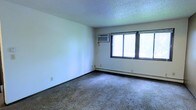 This single-floor space offers a functional and straightforward layout designed for comfortable everyday living. The main living area is finished with wall-to-wall carpeting that adds warmth underfoot, while smooth white walls provide a clean, neutral backdrop throughout. A large, multi-pane window allows generous natural light to pour into the room, creating a bright and open atmosphere. Climate comfort is well-addressed with a wall-mounted air conditioning unit positioned near the window for efficient cooling, complemented by baseboard heating that runs along the perimeter of the room for consistent warmth during cooler months. The textured ceiling adds a subtle architectural detail consistent with the space's practical character. The overall ambiance is understated and move-in ready, offering a blank canvas that can be easily personalized. The open feel of the main room provides flexibility in furniture arrangement, making it adaptable to a variety of uses. This space is well-suited for those seeking a no-frills, functional environment with essential amenities already in place.