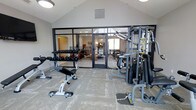 This single-floor fitness space offers a thoughtfully arranged workout environment designed to accommodate a range of exercise needs. The room features vaulted ceilings that lend an open, airy feel, complemented by recessed and flush-mounted lighting that ensures consistent illumination throughout. A large black-framed glass wall and double glass doors create a seamless visual connection to an adjacent lounge or common area, allowing natural light to filter into the space and contributing to a bright, welcoming atmosphere.

The flooring consists of durable, patterned carpet tiles well-suited for a fitness environment. Equipment includes a multi-tiered dumbbell rack stocked with a full range of free weights, an adjustable workout bench, and a sizable multi-function cable and weight stack machine positioned along one wall. A wall-mounted mirror enhances the sense of space while providing visual feedback during workouts. The neutral gray and white color palette, combined with clean lines and modern finishes, gives the fitness area a contemporary aesthetic that feels both functional and polished.