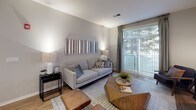 This single-floor residence offers a thoughtfully designed layout with multiple bedrooms, bathrooms, a modern kitchen, and an inviting living area. The living room is anchored by light hardwood-style flooring, neutral walls, and large sliding glass doors that frame lush outdoor greenery, flooding the space with natural light. A geometric wood coffee table and soft area rug add warmth and visual interest.

The bedrooms each carry a distinct character. One features a botanical-print accent wall with integrated sconce lighting, creating a serene retreat. Another showcases a trio of round wall mirrors above a cushioned headboard, complemented by plush carpeting and a modern globe light fixture. A third bedroom includes a wide window with tree views, a rich walnut-toned dresser, and coordinated drapery.

The kitchen is finished with white cabinetry, light countertops, stainless steel appliances, and wood-look flooring. A bold dark accent wall with a wall-mounted wine rack adds a distinctive design touch. Both bathrooms feature tiled tub-shower combinations, warm-toned walls, and wood-style flooring, offering a clean and functional aesthetic throughout.