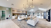 Located at 6857 Poetry Pl in Greenfield, Indiana, this expansive single-story community clubhouse offers a generously sized, open-concept layout designed for both comfort and functionality. The space is defined by its striking vaulted ceilings with large skylights that flood the interior with abundant natural light, complementing the clean white tile flooring found throughout.

The grand entrance welcomes visitors into a bright, airy lobby featuring black-framed glass partition walls, wall-mounted artwork, and a dedicated workstation area. The central living room serves as the heart of the space, anchored by plush white sofas, layered area rugs, and a series of elegant globe and geometric pendant chandeliers. A bold horizontal abstract mural forms a dramatic backdrop, while a wall-mounted TV and built-in bar area with stainless steel appliances add to the amenity-rich environment.

The dining area includes a mix of intimate round tables and a long granite-topped bar with barstool seating, situated beside a large garage-style window that opens to outdoor views. A dedicated game room, visible through black-framed interior windows, houses a foosball table and additional recreation space. Smaller lounge nooks with wall-mounted TVs and café-style seating round out this well-appointed community gathering destination.