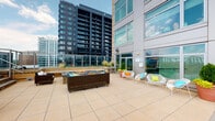 This single-floor space centers around a striking rooftop terrace that offers two distinct outdoor areas designed for relaxation and entertainment. The main terrace is finished with large-format light stone tiles and features a central fire pit surrounded by wicker seating and colorful accent chairs, creating an inviting social gathering space. Glass panel railings frame the perimeter, allowing unobstructed views of the surrounding urban skyline and neighboring high-rise buildings. Lush potted plantings add greenery and texture throughout the terrace. A secondary sun deck area is finished with composite wood decking and outfitted with teak-style chaise lounges positioned to maximize sweeping panoramic views of the cityscape and open sky — ideal for sunbathing or unwinding outdoors. Transitioning indoors, the space includes a half bathroom finished in warm cream-toned walls. It is equipped with a wooden vanity featuring a white countertop sink, a framed mirror, classic chrome fixtures, and brass hardware accents throughout. The bathroom is compact yet functional, with a standard toilet and understated decorative touches. Together, the outdoor terraces and interior bathroom create a well-rounded amenity space suited to both social gatherings and everyday convenience.