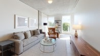 This single-floor unit showcases a thoughtfully designed modern interior defined by a consistent architectural palette of exposed concrete ceilings, light hardwood flooring, and crisp white walls. The open living area is a standout space, featuring large windows that frame lush tree canopy views and flood the room with natural light. A comfortable seating arrangement, glass coffee table, and open metal shelving unit contribute to the space's clean, contemporary aesthetic.

The kitchen is efficiently laid out with warm, flat-front light wood cabinetry extending to the ceiling, white countertops, and a full suite of stainless steel appliances including a gas range, oven, and built-in microwave. A compact dining table with bar-height stools integrates seamlessly into the kitchen area, making efficient use of the space.

The bedroom is a calm retreat with carpet flooring, a natural wood bed frame, wall-mounted reading sconces, a sliding closet, and a window that draws in additional natural light. A full bathroom features a tub-shower combination with a clean, bright finish. Completing the unit's practical offerings is a dedicated in-unit laundry closet with a stacked washer and dryer, conveniently located adjacent to the bathroom vanity area.