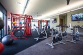 Located at 306 Mezzo Lane in Virginia Beach, Virginia, this well-appointed fitness center offers a comprehensive workout environment across a single open floor. The space is defined by its bold design choices, with striking orange and deep navy accent walls complementing a neutral palette throughout. Floor-to-ceiling glass windows and sliding glass doors flood the interior with natural light while providing direct views of the outdoor pool area and surrounding greenery, creating an energizing atmosphere for exercise.

The cardio zone is well-equipped with multiple treadmills, elliptical trainers, and a recumbent bike, all positioned to take advantage of the expansive outdoor views. A dedicated cycling area features a row of indoor spin bikes, ideal for group or individual cycling sessions. TRX suspension training straps and a red functional training rig add versatility for strength and conditioning workouts, while a stability ball and bench press station round out the free-weight and resistance training options.

Wall-mounted flat-screen televisions are positioned throughout the space for entertainment during workouts. Durable carpet tile flooring covers the workout areas, providing comfort and traction. A ceiling fan ensures air circulation, and the layout allows for free movement between equipment zones. The fitness center combines modern design, ample natural light, and a diverse equipment selection to support a wide range of fitness routines.