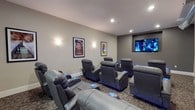 This dedicated movie theater is designed to deliver a premium cinematic experience in a refined, intimate setting. The room is arranged with multiple tiered rows of oversized, dark leather power recliner seats, each equipped with glowing blue accent lights at the base that add a stylish, atmospheric touch. The tiered layout ensures unobstructed sightlines from every seat, maximizing the viewing experience for all guests.

The walls are finished in a neutral gray tone that minimizes glare and keeps the focus on the screen. Framed classic movie posters, including iconic films, are mounted along the walls and accented by soft wall sconces that provide a warm, ambient glow reminiscent of a traditional cinema lobby. Recessed ceiling lighting complements the overall illumination scheme, allowing for adjustable brightness to suit any screening.

A large flat-panel display screen is mounted prominently at the front of the room, serving as the focal point of the theater. Carpeted flooring in a subtle pattern adds to the acoustic comfort and classic movie-house aesthetic of the space. Overall, this theater combines comfort, style, and functionality to create an inviting environment ideal for private screenings and entertainment gatherings.
