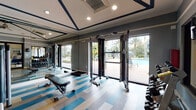 Located at 10200 Falcon Pine Blvd in Orlando, Florida, this well-appointed fitness center offers a comprehensive workout environment designed for both strength training and functional fitness. The single-floor space features a striking multi-toned vinyl tile floor in shades of blue, tan, and white, adding a vibrant, modern aesthetic throughout. The room is equipped with a cable crossover machine, a full rack of dumbbells, multiple weight benches including adjustable Matrix-brand benches, and a variety of selectorized strength machines. Large floor-to-ceiling windows and glass doors line one wall, flooding the space with abundant natural light and providing direct views of a resort-style swimming pool and swaying palm trees beyond — creating an energizing, Florida-inspired atmosphere. Full-length mirrors along the walls enhance the sense of openness and allow users to monitor their form. A coffered ceiling with recessed lighting and a ceiling fan adds a refined architectural touch overhead. A glass-walled room adjacent to the main floor, featuring warm hardwood flooring, appears to serve as a dedicated yoga or group fitness studio. Access to a broader amenity space — including what appears to be a lounge and billiards area — is visible through glass-paned double doors, connecting the fitness center to the larger community facility.