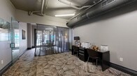 This 340-square-foot business center is thoughtfully designed across a single floor, offering a blend of functional and collaborative spaces suited for professional use. The main area features an industrial-modern aesthetic, with exposed ductwork running along the ceiling, bold geometric-patterned carpeting, and neutral gray walls that create a clean, contemporary atmosphere. Fluorescent linear lighting ensures a well-lit environment throughout.

A built-in workstation along one wall provides dual desk spaces equipped with a printer and resident Wi-Fi access, accompanied by dark ergonomic chairs — ideal for individual work or administrative tasks. Decorative touches, including a sculptural gold accent piece and a table lamp, add warmth to the practical setup.

A semi-private conference room is separated from the main area by sheer gray curtains, offering a natural wood live-edge conference table, mesh task chairs, a wall-mounted flat-screen TV, and a stylish multi-bulb brass chandelier. A sideboard with decorative lamps and sculptures completes the refined look, while large glass doors allow natural light to filter in.

An additional quiet nook features a single lounge chair and two framed abstract wall art pieces, providing a calm retreat for reading or reflection. Dark gray interior doors and consistent carpeting unify the overall design of this versatile amenity space.