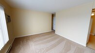 This single-floor space presents a clean and straightforward layout designed for functional living. The main room is finished with wall-to-wall beige carpeting that provides a warm and comfortable underfoot feel, complementing the soft yellow-painted walls that lend the space a bright, neutral ambiance. White baseboards and trim create a crisp contrast against the walls, adding a sense of polish to the overall aesthetic. The standard ceiling height contributes to a practical, well-proportioned feel throughout the room. A white interior door is visible toward the rear of the space, indicating a connection to an additional room or hallway beyond, suggesting a multi-room floor plan. Electrical outlets are positioned along the walls, offering convenient access throughout the room. The space is unfurnished, allowing for a clear view of the architectural layout and providing flexibility for a variety of uses and configurations. The neutral color palette and clean finishes throughout make this space highly adaptable, offering a blank canvas that can be personalized to suit a range of preferences and needs. Overall, the space conveys a sense of simplicity and practicality with a welcoming, light-toned interior.