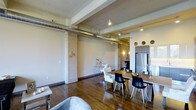 Located in Dallas, this urban loft showcases a compelling blend of industrial character and contemporary design. The open-concept layout flows seamlessly from the living area through to the dining and kitchen spaces, creating a sense of expansive, connected living. Rich hardwood floors run throughout, grounding the space with warmth and texture.

Architectural details are a defining feature here. Exposed concrete columns, visible overhead ductwork, and structural ceiling beams lend a raw, industrial aesthetic that is balanced by modern finishes and thoughtful furnishings. The elevated ceilings amplify the sense of openness and volume throughout the space.

The dining area is anchored by a substantial wood table surrounded by a mix of modern black and white chairs, creating a striking visual contrast. Nearby, the kitchen features sleek cabinetry and stainless steel appliances, including a full-sized refrigerator, offering a clean and functional counterpoint to the industrial surroundings. Track lighting and accent fixtures provide layered illumination throughout, complementing the open layout.

The living area is partially visible and hints at a comfortable, curated setup. Overall, the space delivers a sophisticated urban atmosphere with an engaging mix of raw architectural elements and modern design sensibilities.
