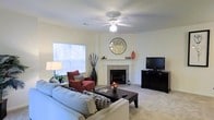 This single-floor space centers around a welcoming living room designed for comfort and functionality. The room is finished with wall-to-wall carpeting in a neutral tone, contributing to a warm and inviting atmosphere. A tile-surround fireplace serves as the focal point of the space, flanked by a decorative mantel that adds a classic architectural touch. Above, a ceiling fan with an integrated light fixture provides both air circulation and ambient lighting throughout the room. A generously sized window fitted with blinds allows natural light to fill the space while offering a view of the surrounding greenery outdoors. The walls are painted in a soft, neutral hue that complements the light carpeting and creates a bright, airy feel. The layout accommodates a comfortable seating arrangement, with ample room for a sofa, accent chair, and a central coffee table. Additional space along one wall provides an ideal area for an entertainment setup. The overall ambiance is relaxed and livable, with a straightforward design that balances practicality with everyday comfort, making it a functional and pleasant living environment.