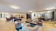 This expansive two-floor space offers a thoughtfully designed environment that blends modern aesthetics with functional communal living. The main floor serves as the social hub, featuring an open-plan living and lounge area finished with wide-plank light oak hardwood flooring that runs throughout. Multiple seating arrangements — outfitted with plush, sculptural furniture in muted earth tones — create inviting conversation areas suited for both casual gatherings and focused collaboration. Exposed concrete columns and textured dark brick accent walls provide an industrial contrast to the clean white ceilings and warm wood tones.

A glass-walled private room with a sculptural chandelier adds a refined, semi-enclosed gathering option, while a dedicated dining area offers a long communal table with gold-accented bar stools set against a raw concrete wall. The kitchen is seamlessly integrated nearby. A formal meeting room features a large conference table, leather seating, and dramatic pendant lighting.

The upper floor opens onto a generous mezzanine-level lounge bathed in natural light from floor-to-ceiling windows overlooking lush greenery. A sleek metal railing frames the open stairwell connecting both levels. Additional seating alcoves with olive-toned velvet chairs offer quieter retreats throughout the space, completing a well-balanced and visually striking interior.
