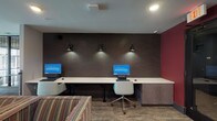 Located at 12 W Gay St in the heart of downtown Columbus, Ohio, this business center offers a focused and professional environment designed to meet the needs of productivity-minded users. The space is thoughtfully laid out on a single floor, providing a compact yet fully functional setting for work and connectivity. A long, continuous white desk runs along a striking dark brick accent wall, accommodating multiple computer workstations equipped with monitors and keyboards. Sleek wall-mounted directional lamps positioned above each workstation cast focused illumination across the workspace, complementing the recessed ceiling lighting overhead. The accent wall's deep, dark tone creates a strong visual contrast against the crisp white desk surface, lending the space a modern and polished aesthetic. A bold burgundy wall adds a pop of color to the room, contributing to the overall contemporary design. Carpeted flooring in a neutral tone grounds the space, while an ergonomic chair at the workstation ensures user comfort. Built-in cabinetry provides additional storage, keeping the area organized and efficient. The overall ambiance is clean, professional, and well-appointed, making this business center a practical and inviting destination for guests in the Columbus area.