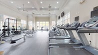 This single-floor fitness center is designed to accommodate a wide range of exercise needs within two distinct but connected zones. The main cardio area is an expansive, light-filled space distinguished by its soaring coffered ceilings adorned with crown molding, ceiling fans, and recessed lighting. Large white-framed windows line the walls, inviting generous natural light and offering views of the surrounding exterior. A row of treadmills, ellipticals, and a stair climber are positioned throughout, each equipped with individual monitors. Wall-mounted flat-screen televisions provide additional entertainment options throughout the space.

The strength training zone features durable rubber flooring and is lined with full-length mirrors that amplify the sense of space. This area houses a comprehensive selection of cable machines, a rack with free weights and barbells, adjustable benches, and rowing machines, offering a thorough range of resistance training options. Stationary bikes are also available for low-impact cardio.

The fitness center is accessed through elegant glass-paneled French doors, reinforcing the refined aesthetic carried throughout. The overall ambiance is clean, bright, and professional, reflecting a thoughtfully designed facility suited for both casual and dedicated fitness routines.