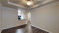 This single-floor residence in Dallas offers a thoughtfully designed open-concept layout that seamlessly connects the living and kitchen areas. The living room makes a strong first impression with its elegant tray ceiling adorned with crown molding and a ceiling fan, adding both architectural interest and functional comfort. Rich, dark hardwood-style flooring runs throughout, providing a warm and polished foundation that contrasts beautifully with the neutral, light-toned walls.

The kitchen is open to the living area via a breakfast bar counter finished with granite countertops, creating an ideal space for casual dining or entertaining. White cabinetry lines the kitchen walls, complemented by modern appliances including a range and microwave, while a sink sits beneath adequate lighting. The pass-through design enhances the sense of openness and allows natural interaction between spaces.

Crown molding details are carried consistently throughout the space, lending a refined, cohesive character to the interior. A hallway visible from the main living area leads to additional rooms, suggesting a practical and well-organized floor plan. Overall, the space combines classic architectural detailing with contemporary finishes to create a clean, inviting atmosphere suited for comfortable everyday living in Dallas.