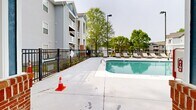 Located at 4900 Chandler Ridge Cir in Raleigh, NC, this outdoor amenity area centers around a large, rectangular swimming pool with clear turquoise water. The pool is equipped with stainless steel entry rails and is designated as a no-diving pool, reflecting a safe and well-maintained environment. Surrounding the pool is an expansive concrete sun deck that comfortably accommodates numerous lounge chairs and chaise loungers, ideal for relaxing in the North Carolina sunshine. A white pergola structure on one side of the deck provides a shaded seating area, offering a cooler retreat for residents. The entire pool area is enclosed by a black iron security fence, ensuring privacy and safety. Adjacent to the pool sits a single-story clubhouse building with brick accent columns, a pitched shingle roof, and a prominent chimney. The building features a covered patio area with direct pool-side access, accented by bright blue entry doors that add a pop of color to the facade. Mature trees line the perimeter of the space, providing natural greenery and partial shade. The surrounding multi-story residential buildings are visible beyond the fencing, situating this amenity space firmly within a well-established residential community.