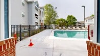 Located at 4900 Chandler Ridge Cir in Raleigh, NC, this outdoor amenity area centers around a large, rectangular swimming pool with clear turquoise water. The pool is equipped with stainless steel entry rails and is designated as a no-diving pool, reflecting a safe and well-maintained environment. Surrounding the pool is an expansive concrete sun deck that comfortably accommodates numerous lounge chairs and chaise loungers, ideal for relaxing in the North Carolina sunshine. A white pergola structure on one side of the deck provides a shaded seating area, offering a cooler retreat for residents. The entire pool area is enclosed by a black iron security fence, ensuring privacy and safety. Adjacent to the pool sits a single-story clubhouse building with brick accent columns, a pitched shingle roof, and a prominent chimney. The building features a covered patio area with direct pool-side access, accented by bright blue entry doors that add a pop of color to the facade. Mature trees line the perimeter of the space, providing natural greenery and partial shade. The surrounding multi-story residential buildings are visible beyond the fencing, situating this amenity space firmly within a well-established residential community.