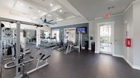 Located at 3306 S Fry Rd in Katy, Texas, this well-appointed fitness center offers a comprehensive workout environment across a single floor. The space is organized into distinct functional zones, each designed to support a variety of exercise routines.

The main workout area is outfitted with a lineup of cardio machines including treadmills and ellipticals, complemented by a selection of strength training equipment such as cable machines and a dumbbell rack. Full-length mirrors along the walls enhance the sense of space and allow users to monitor their form. A mounted flat-screen television provides entertainment during workouts, and a large ceiling fan ensures comfortable air circulation throughout.

A dedicated cycling studio houses multiple stationary spin bikes arranged for group or individual sessions, with windows allowing natural light to filter in. Adjacent to the cycling area, a functional training zone offers stability balls, resistance bands, and medicine balls neatly organized on a rack — ideal for stretching, core work, and cross-training.

The facility features durable rubber flooring throughout, crown molding, recessed lighting, and a neutral gray color palette that gives the space a clean, polished appearance. Windows fitted with blinds bring in natural light while maintaining privacy. Restroom access is available on-site, rounding out the amenities of this fully equipped community fitness center.