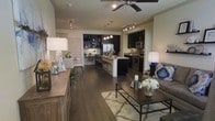 This thoughtfully designed single-floor residence showcases an open-concept layout that effortlessly connects the living, dining, and kitchen areas into one cohesive and functional space. Rich, dark hardwood flooring flows throughout, grounding the interior with warmth and sophistication.

The gourmet kitchen is a standout feature, outfitted with stainless steel appliances including a refrigerator, microwave, and range, complemented by sleek dark cabinetry and a contrasting light-toned island. The island doubles as a breakfast bar with seating for multiple guests, making it ideal for casual dining and entertaining. Pendant lighting above the island adds a refined touch, while track lighting throughout ensures the space is well-illuminated.

The living area is generously proportioned, centered around a low-profile coffee table and plush seating. A ceiling fan overhead provides both comfort and a design element. Neutral walls allow the darker furnishings and flooring to take center stage. A sideboard along one wall provides additional storage and display space.

A dedicated workspace or reading nook is visible in the background, offering a quiet retreat within the open floor plan. The overall ambiance is modern and inviting, blending functionality with contemporary style in a cohesive, well-appointed interior.