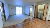 This single-floor residential space offers a functional and straightforward layout designed for comfortable everyday living. The main living area is a standout feature, combining warm hardwood flooring with plush wall-to-wall carpeting, creating a clear and natural division between the entry and lounge zones. Large sliding glass doors flood the room with abundant natural light, lending an open and airy atmosphere to the interior. Vertical blinds provide privacy and light control as needed. A wall-mounted air conditioning unit and baseboard heating ensure year-round climate comfort throughout the space. The textured ceiling adds a classic residential character to the room. An open doorway leads to an additional room, also finished with carpeting and a window, offering flexibility for use as a bedroom or secondary living space. A closet with a sliding door is conveniently positioned nearby, providing practical storage. The overall palette is neutral and clean, with white walls and warm wood trim framing each space. The layout is efficient and accessible, making it well-suited for a range of living arrangements.
