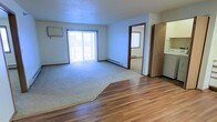 This single-floor residential space offers a functional and straightforward layout designed for comfortable everyday living. The main living area is a standout feature, combining warm hardwood flooring with plush wall-to-wall carpeting, creating a clear and natural division between the entry and lounge zones. Large sliding glass doors flood the room with abundant natural light, lending an open and airy atmosphere to the interior. Vertical blinds provide privacy and light control as needed. A wall-mounted air conditioning unit and baseboard heating ensure year-round climate comfort throughout the space. The textured ceiling adds a classic residential character to the room. An open doorway leads to an additional room, also finished with carpeting and a window, offering flexibility for use as a bedroom or secondary living space. A closet with a sliding door is conveniently positioned nearby, providing practical storage. The overall palette is neutral and clean, with white walls and warm wood trim framing each space. The layout is efficient and accessible, making it well-suited for a range of living arrangements.