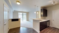This single-floor residence offers a well-designed, open-concept layout that seamlessly connects the kitchen and living areas. The kitchen is equipped with stainless steel appliances, including a range, microwave, and refrigerator, complemented by dark espresso cabinetry, white countertops, and a stainless steel sink. A peninsula with pendant lighting provides additional workspace and casual dining options. Hardwood-style flooring runs through the kitchen and entry areas, transitioning to plush carpeting in the living and bedroom spaces.

The living area is generously sized, featuring a sliding glass door that opens to a private balcony with outdoor views, allowing ample natural light to fill the space. The entry area includes track lighting and a defined transition between the wood and carpeted zones.

The bedrooms are spacious and carpeted, each featuring ceiling fans and vertical blinds over windows that bring in natural light. One bedroom includes a baseboard heater, offering individual climate control.

The bathroom features a dark espresso vanity with white countertops, a large mirror with overhead lighting, wood-style flooring, and access to an open shelving closet area. The overall aesthetic throughout is clean, neutral, and functional.
