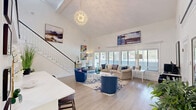 Located at 201 Treybrooke Dr in Morrisville, NC, this two-story space offers a well-appointed layout spread across two distinct levels. The main floor serves as the primary gathering and lounge area, featuring soaring vaulted ceilings that create an open, airy atmosphere. Light wood flooring flows throughout, complemented by large windows and glass French doors that flood the space with natural light and provide views of the surrounding greenery. The main level includes comfortable seating areas adorned with colorful contemporary artwork, as well as a dedicated computer workstation with multiple monitors and bar-height seating — ideal for residents or visitors needing a productive workspace. Clean white walls and carefully chosen décor give the area a bright, welcoming ambiance. The upper level, accessible via an interior staircase with an open mezzanine railing, offers a more private and versatile setting. Carpeted flooring, vaulted ceilings with angled rooflines, and generous natural light from high clerestory and standard windows define this floor. A formal conference table with ample seating anchors one end of the upper level, making it well-suited for meetings or group gatherings, while an adjacent open lounge area provides additional flexible space for smaller gatherings or quiet retreats.