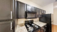 Located at 9449 Dayton Pike in Soddy-Daisy, Tennessee, this single-floor unit offers a well-designed layout with consistent finishes throughout. Warm hardwood-style flooring flows seamlessly across the main living areas and hallways, creating a cohesive and inviting atmosphere. The living area is open and well-lit, featuring a door that leads to an outdoor space, allowing natural light to fill the room. A pass-through bar with granite-style countertops connects the living area to the kitchen, enhancing the open-concept feel. The kitchen is equipped with dark espresso-toned cabinetry featuring glass-panel upper doors and brushed nickel hardware, complemented by granite-style countertops. Black appliances, including a coil-burner range, over-the-range microwave, and refrigerator, complete the cooking space. The bathroom mirrors the kitchen's design aesthetic, with matching dark cabinetry, granite-style countertops, a single-basin sink, a large mirror, and a shower-tub combination. A generously sized walk-in closet with wire shelving and hanging rods provides ample storage. Neutral, warm-toned walls throughout give the space a clean and move-in-ready appearance.