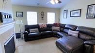 This single-floor home offers a warm and inviting layout designed for comfortable everyday living. The living room serves as the heart of the space, centered around a generously sized dark leather sectional sofa with reclining features, providing ample seating for family and guests alike. Soft, neutral-toned carpeting runs underfoot, complementing the light beige walls that give the room a bright and airy feel. Multiple windows allow natural light to filter in, enhancing the open atmosphere throughout the day. A ceiling fan with an integrated light fixture is mounted overhead, offering both practical ventilation and additional illumination. Wall décor, including framed artwork and a decorative wall clock, adds a personalized touch to the living area. The overall ambiance of the home is relaxed and functional, with a layout that prioritizes comfort and livability. The single-floor design ensures easy accessibility throughout, making it a practical and welcoming space suited for a variety of lifestyles.