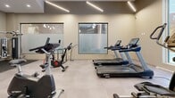 This state-of-the-art fitness center, spanning 910 square feet, offers a comprehensive workout experience in a contemporary setting. The space is thoughtfully designed with a focus on functionality and aesthetics. Large windows flood the area with natural light, creating an inviting and energizing atmosphere. The layout is well-organized, featuring distinct zones for cardio and strength training. 

The cardio section is equipped with a variety of machines, including treadmills, stationary bikes, and ellipticals, all facing the expansive windows to provide an engaging view during workouts. The strength training area boasts a selection of free weights and resistance machines, catering to diverse fitness needs.

Architectural elements such as exposed ceilings with modern lighting fixtures and sleek structural columns contribute to the space's contemporary feel. The flooring is a durable, low-maintenance material suitable for high-traffic gym environments. Built-in storage solutions and wall-mounted amenities, like sanitizer dispensers and towel holders, maximize usable floor space.

The gym's design prioritizes cleanliness and organization, with ample room between equipment for safe navigation and social distancing. The neutral color palette, accented by the occasional pop of color from equipment or storage units, creates a calm and focused environment conducive to effective workouts.