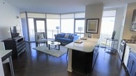 This elegant single-floor residence seamlessly blends modern design with comfort, offering an open-concept layout that maximizes both space and natural light. The living area is anchored by expansive floor-to-ceiling windows that wrap around the corner of the room, flooding the interior with daylight and providing sweeping outdoor views through roller shade window treatments. Rich, dark hardwood flooring runs throughout, creating a striking contrast against the bright, airy atmosphere.

The living space is thoughtfully designed, featuring a central seating arrangement with a classic blue sofa, accent chairs, a glass-top coffee table, and a plush area rug that defines the lounge zone. Curated artwork adorns the walls, adding a refined, gallery-like touch to the space.

Flowing directly from the living area is the kitchen, highlighted by a substantial dark-cabinetry island topped with a crisp white countertop surface. The island serves as both a functional workspace and a natural divider between the kitchen and living areas, maintaining the open, connected feel of the floor plan.

The space also benefits from access to an outdoor balcony, visible through the glass doors, further enhancing the sense of openness and bringing the outside in. Overall, the residence offers a sophisticated and welcoming environment defined by clean lines, quality finishes, and abundant natural light.