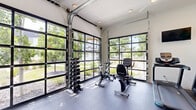 Located in Spokane Valley, WA, this contemporary fitness center offers a well-equipped workout space designed with both functionality and aesthetics in mind. The facility is defined by its striking floor-to-ceiling glass panel garage doors, which flood the interior with natural light and provide open views of the lush, tree-lined surroundings. The durable rubber flooring throughout provides a safe and practical surface for a wide range of exercises. The cardio zone is well-appointed with True brand treadmills, ellipticals, a spin bike, and a recumbent bike, complemented by wall-mounted flat-screen TVs. A dedicated strength and free weights area features a tiered dumbbell rack and a cable resistance machine, catering to varied training needs. The functional training section includes a full wall mirror, stability balls in multiple sizes, a workout bench, and instructional fitness posters. Exposed industrial-style ductwork along the ceiling adds a modern design element, while ceiling fans ensure comfortable airflow. Recessed lighting keeps the space well-lit throughout. The overall ambiance is bright, open, and inviting, making this fitness center a comprehensive and appealing amenity.