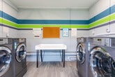Located at 1800 Kuser Rd in Trenton, New Jersey, this single-story shared laundry facility is designed for functionality and ease of use. The space is defined by its painted concrete block walls, decorated with bold horizontal stripes in teal, green, and gray, giving the room a clean and lively aesthetic.

The facility is equipped with multiple commercial-grade LG front-loading washing machines, arranged along the perimeter walls to maximize usable floor space. A central folding table with a modern hairpin-leg base provides a convenient surface for sorting and folding laundry. An information board mounted on the wall displays notices and payment instructions, including mobile payment options.

Light wood-look flooring runs throughout, contributing to a fresh and well-maintained appearance. Natural light enters through multiple windows, including a side window and a glazed exterior door, offering views of the surrounding greenery. Exposed ventilation ductwork runs along the upper walls, reflecting the utilitarian nature of the space. A waste bin is conveniently positioned for resident use. The overall layout is efficient and accessible, serving as a practical shared amenity for the surrounding community.