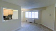 This single-floor residence offers a clean, functional layout designed for comfortable everyday living. The living area is finished with plush wall-to-wall carpeting and neutral, light-toned walls that create a bright and welcoming atmosphere. A wide, multi-pane window fitted with horizontal blinds allows abundant natural light to fill the space, enhancing its open and airy feel. A wall-mounted HVAC unit is conveniently positioned beneath the window, ensuring year-round climate control. A notable architectural feature is the pass-through window connecting the kitchen to the living area, which promotes an open-concept feel and allows for easy interaction between the two spaces. The kitchen, visible through the pass-through, features cabinetry and is well-integrated into the overall layout. A pendant light fixture hangs at the transition point between the kitchen and living areas, providing focused overhead lighting. White baseboards and trim throughout the space offer a crisp contrast to the soft carpeting and walls. The textured ceiling adds subtle visual interest. Overall, the space presents a practical and inviting environment with a cohesive, understated design aesthetic suited to a variety of lifestyles.