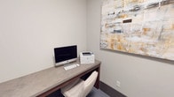 This single-floor workspace is a compact, purpose-built office area designed for productivity and functional use. The room is finished with neutral, warm-gray walls that create a clean and professional atmosphere. Carpeted flooring in a subtle blue-gray tone adds a layer of comfort underfoot throughout the space.

The primary workstation consists of a built-in desk with a stone-look composite surface, supported by warm wood-toned cabinetry below. The desk accommodates a full-sized iMac desktop computer alongside an HP laser printer, making the space fully equipped for both digital and print tasks. A cushioned white task chair completes the workstation setup.

A standout decorative element is a large abstract canvas artwork displayed prominently on the wall above the desk. The painting features a layered composition in gold, white, gray, and charcoal tones, introducing visual warmth and a contemporary aesthetic to an otherwise minimal space.

A black steel-framed glass door serves as the primary entry point, offering transparency into an adjacent common or collaborative area. The glazed sidelight panel beside the door further enhances visual connectivity between spaces while maintaining a modern, industrial-chic design language. A matte black door lever hardware detail reinforces the cohesive design palette throughout the space.