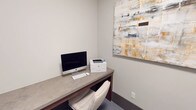 This single-floor workspace is a compact, purpose-built office area designed for productivity and functional use. The room is finished with neutral, warm-gray walls that create a clean and professional atmosphere. Carpeted flooring in a subtle blue-gray tone adds a layer of comfort underfoot throughout the space.

The primary workstation consists of a built-in desk with a stone-look composite surface, supported by warm wood-toned cabinetry below. The desk accommodates a full-sized iMac desktop computer alongside an HP laser printer, making the space fully equipped for both digital and print tasks. A cushioned white task chair completes the workstation setup.

A standout decorative element is a large abstract canvas artwork displayed prominently on the wall above the desk. The painting features a layered composition in gold, white, gray, and charcoal tones, introducing visual warmth and a contemporary aesthetic to an otherwise minimal space.

A black steel-framed glass door serves as the primary entry point, offering transparency into an adjacent common or collaborative area. The glazed sidelight panel beside the door further enhances visual connectivity between spaces while maintaining a modern, industrial-chic design language. A matte black door lever hardware detail reinforces the cohesive design palette throughout the space.
