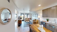 This thoughtfully designed single-floor residence offers a seamless blend of modern aesthetics and functional living. The open-concept layout connects the living, dining, and kitchen areas into one cohesive space, bathed in natural light from large black-framed windows and a glass door that opens to the outdoors. Light-toned hardwood flooring flows throughout, enhancing the bright, airy atmosphere.

The living area is generously proportioned, anchored by a plush sectional sofa and complemented by a gallery-style arrangement of framed artwork on crisp white walls. The adjoining dining space features a rustic wood table, providing a warm contrast to the contemporary surroundings.

The kitchen is outfitted with floor-to-ceiling flat-front cabinetry in a natural wood-grain finish, stainless steel appliances including a French door refrigerator, a range with over-the-range microwave, and clean white countertops that offer ample prep space.

The bedroom is a restful retreat, featuring large black-framed windows that frame views of the surrounding neighborhood and fill the room with natural light. Soft carpeting and warm wood furnishings complete the comfortable atmosphere.

The bathroom is clean and modern, with light wood vanity cabinetry, a full tub-shower combination, and recessed lighting, maintaining the consistent contemporary design language found throughout the residence.