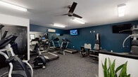 Located at 307 Simmons St in New Bern, North Carolina, this single-story fitness center offers a spacious, well-organized workout environment designed to accommodate a range of exercise needs. The facility is laid out across an open floor plan with distinct zones for cardio, strength training, and free weights, all unified by durable, dark patterned carpet flooring throughout.

The cardio area is equipped with multiple treadmills, elliptical trainers, a stationary bike, and a rowing machine, complemented by wall-mounted televisions for entertainment during workouts. A large mirrored wall along one side of the room enhances the sense of space and allows users to monitor their form. The strength training section features a multi-station cable and weight machine, a leg extension bench, and a dedicated dumbbell rack with an assortment of free weights.

The space is kept comfortable by multiple ceiling fans and wall-mounted mini-split HVAC units. Fluorescent lighting provides bright, even illumination throughout. Natural light filters in through several windows, and double glass entry doors contribute additional brightness near the entrance. Decorative potted plants are placed throughout, adding a welcoming touch to the utilitarian environment. A water fountain and an additional private room are also accessible within the facility.