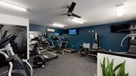 Located at 307 Simmons St in New Bern, North Carolina, this single-story fitness center offers a spacious, well-organized workout environment designed to accommodate a range of exercise needs. The facility is laid out across an open floor plan with distinct zones for cardio, strength training, and free weights, all unified by durable, dark patterned carpet flooring throughout.

The cardio area is equipped with multiple treadmills, elliptical trainers, a stationary bike, and a rowing machine, complemented by wall-mounted televisions for entertainment during workouts. A large mirrored wall along one side of the room enhances the sense of space and allows users to monitor their form. The strength training section features a multi-station cable and weight machine, a leg extension bench, and a dedicated dumbbell rack with an assortment of free weights.

The space is kept comfortable by multiple ceiling fans and wall-mounted mini-split HVAC units. Fluorescent lighting provides bright, even illumination throughout. Natural light filters in through several windows, and double glass entry doors contribute additional brightness near the entrance. Decorative potted plants are placed throughout, adding a welcoming touch to the utilitarian environment. A water fountain and an additional private room are also accessible within the facility.
