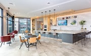 Located on Schering Drive in Union, NJ, this single-floor amenity space combines contemporary design with high-end finishes throughout. The space is anchored by a showpiece kitchen outfitted with stainless steel double wall ovens, a professional-grade gas range, and an expansive stone-topped island with bar seating. Light wood cabinetry, a textured tile backsplash, open display shelving, and an integrated flat-screen TV give the kitchen both functionality and visual interest. Large-format neutral tile flooring extends throughout, maintaining a cohesive aesthetic across the entire space.

The lounge and dining areas are defined by floor-to-ceiling glass curtain walls that flood the interior with natural light and offer sweeping views of the surrounding development. Bold red upholstered chairs paired with round bistro tables create a vibrant social atmosphere, while decorative latticed columns add architectural texture. A dramatic central atrium features a large skylight overhead and a sculptural chandelier suspended above glass-railed mezzanine walkways and open staircases, lending the space a striking sense of vertical grandeur and openness.