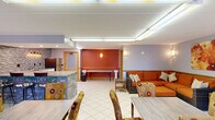 Located on Berkshire Lane in Burton, Ohio, this single-floor community space offers a well-appointed and functional layout across three distinct areas. The open main room serves as the heart of the space, combining a comfortable lounge and dining area beneath a drop ceiling with recessed cove lighting. The lounge is furnished with a wicker-frame sectional sofa in bold orange upholstery, accent chairs with floral patterning, and wicker side tables — all accented by wall-mounted sconces that provide warm ambient lighting. A vibrant orange accent wall anchors the dining nook, where a folding table and decorative pedestals create a flexible event setup. The kitchenette is a standout feature, showcasing white shaker cabinetry, granite-look countertops, a stainless steel refrigerator, microwave, and a striking mosaic tile backsplash illuminated by an industrial-style vanity light. Large vertical blinds throughout admit natural light while maintaining privacy. The restroom features classic beadboard wainscoting and clean, neutral finishes. Wall-unit HVAC ensures climate control throughout. White tile flooring with warm wood trim unifies the space, creating a cohesive, welcoming environment suited for community events, meetings, or social gatherings.