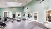 Located at 32 Ashbury Woods Dr in Huntsville, Alabama, this fitness center is a thoughtfully designed workout space that combines architectural character with a comprehensive range of exercise equipment. The room is defined by its striking vaulted ceilings, which lend an open, airy feel, and a large skylight that floods the space with natural light. Soft sage-green walls contribute to a calm and energizing atmosphere throughout.

Large arched windows fitted with white blinds line the walls, allowing additional natural light and views of the surrounding greenery. A full-length wall mirror visually expands the space and supports proper form during workouts. Ceiling fans ensure comfortable airflow during intense exercise sessions.

The fitness floor is finished with durable rubber flooring suited for high-traffic activity. Equipment includes multiple treadmills and elliptical machines from True Fitness, a multi-station cable strength system, a weight bench, a rack of dumbbells, and medicine balls. A wall-mounted flat-screen television provides entertainment during workouts.

Multiple French doors offer access to the exterior, enhancing ventilation and connection to the outdoors. Recessed lighting complements the natural light to keep the space bright at all hours. The overall design balances functionality with an inviting aesthetic, making this fitness center a well-appointed facility for a full range of cardio and strength training activities.