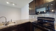 Located at 5451 Olivia Michal Pl in Westerville, Ohio, this single-floor unit offers a functional and well-appointed layout designed for comfortable everyday living. The open-concept living and kitchen area serves as the heart of the home, where pendant lighting illuminates a peninsula-style counter with granite-look laminate surfaces. The kitchen is outfitted with dark espresso cabinetry, a mosaic tile backsplash, black appliances including an electric range and over-the-range microwave, and a stainless steel sink. Open shelving adds a decorative accent to the cabinetry design. The living area features soft wall-to-wall carpeting and a glass-paneled door that invites natural light and provides outdoor access. The bedroom is a comfortable retreat, also finished with plush carpeting, neutral tones, a bold accent wall, and access to a walk-in closet. The bathroom features warm-toned luxury vinyl plank flooring, a white vanity with a clean countertop, and a neutral color palette. A dedicated in-unit laundry area accommodates a full-sized washer and dryer with overhead shelving for additional storage. Throughout the unit, clean white trim, soft neutral wall tones, and consistent finishes create a cohesive and inviting atmosphere.